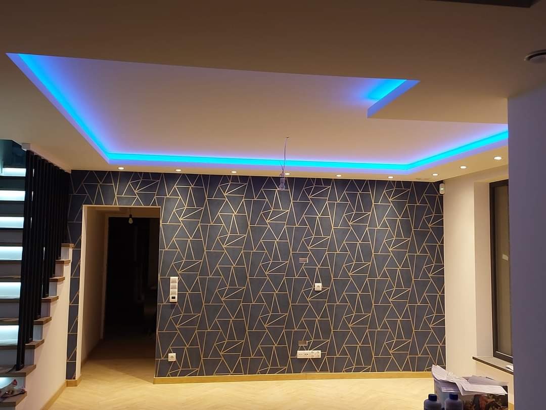 Nowoczesne wnętrze z podwieszanym sufitem z niebieskim oświetleniem LED, ściana pokryta tapetą w geometryczne wzory, widoczne schody i fragment okna.