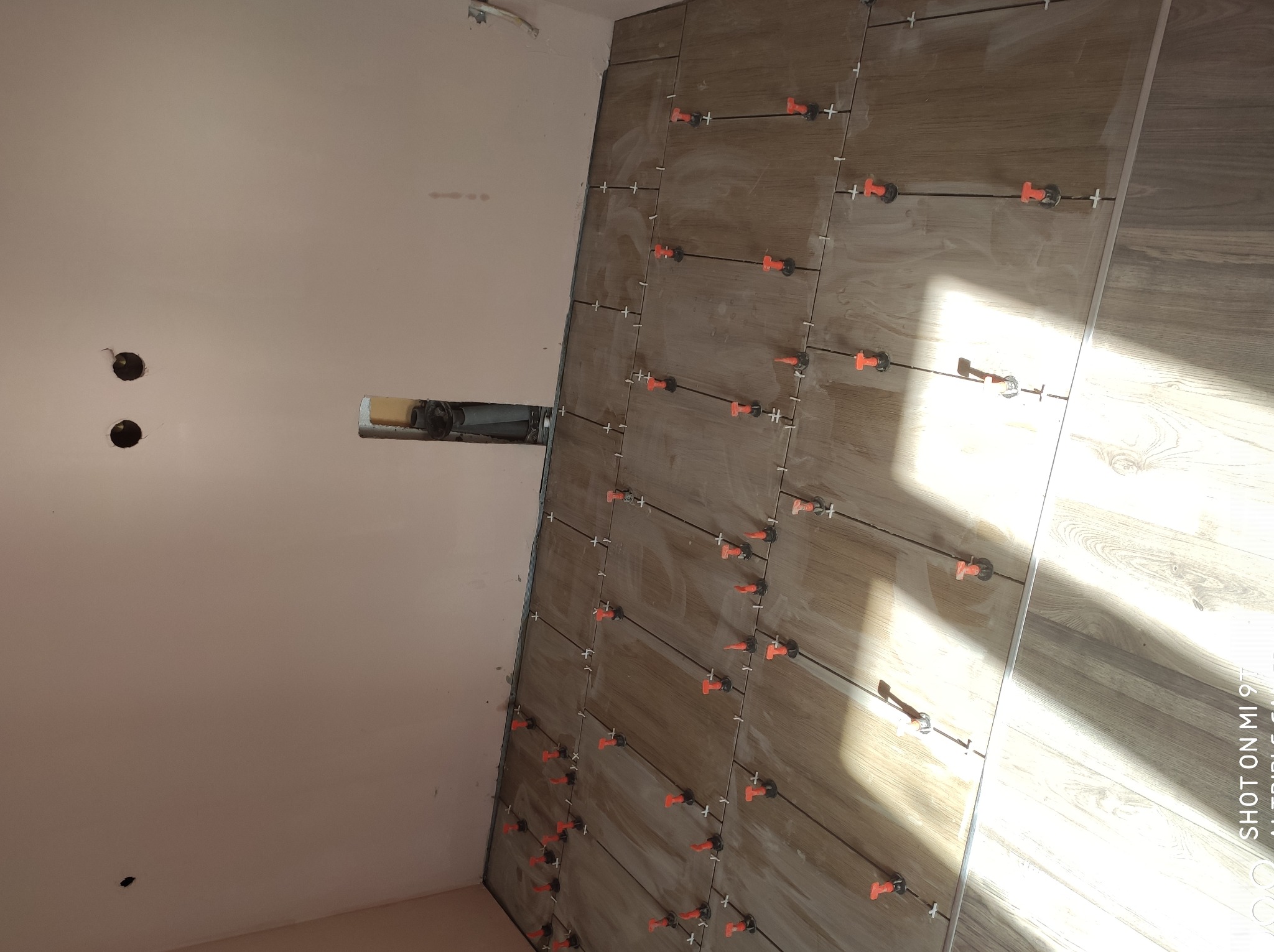 Montaż płytek imitujących drewno na ścianie, z użyciem pomarańczowych klipsów poziomujących, widoczne otwory w ścianie obok, instalacja w trakcie realizacji.