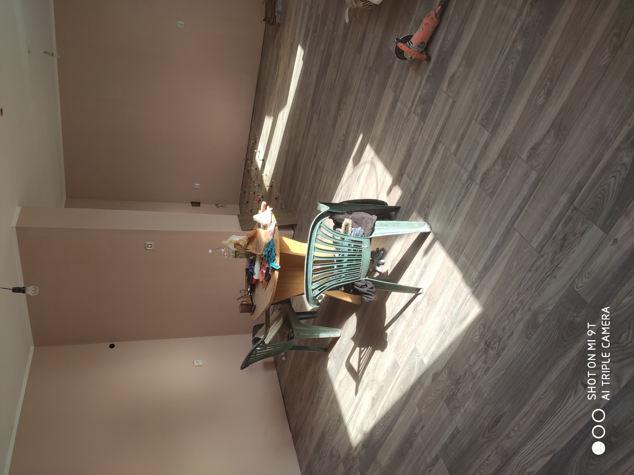 Przygotowania do remontu: pokój z drewnopodobną podłogą w trakcie układania, widoczne narzędzia, stół z akcesoriami i plastikowe krzesła.