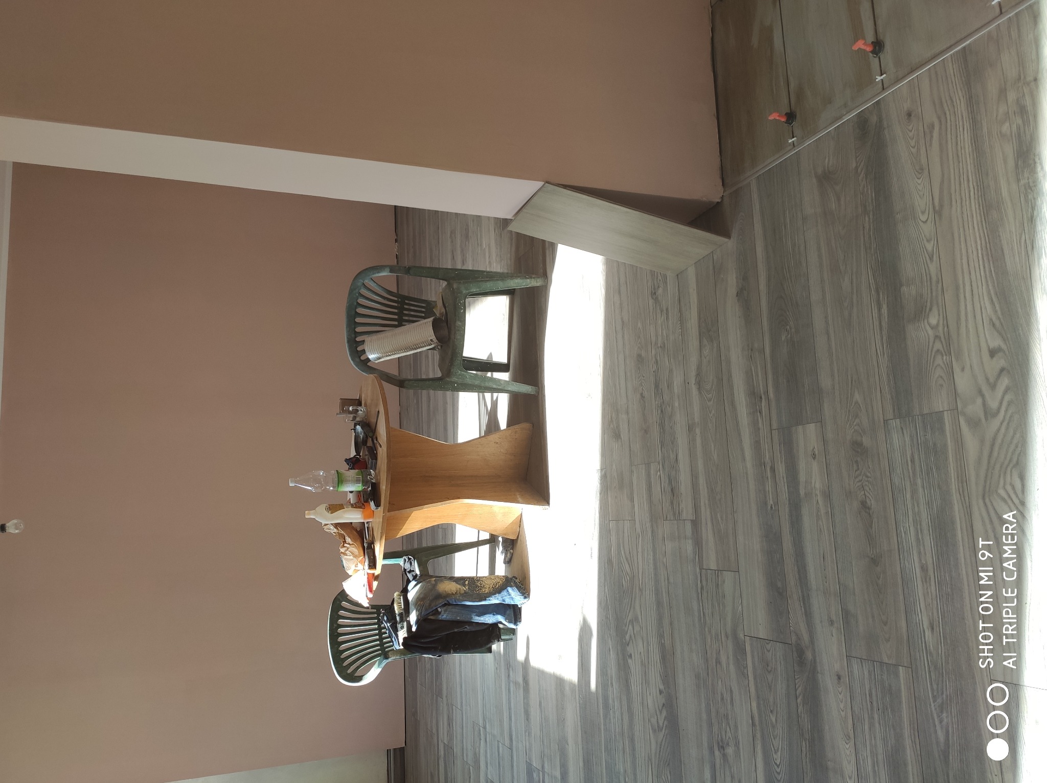 Układanie paneli podłogowych z widocznymi plastikowymi dystansami przy ścianie, stolik z narzędziami i pustą puszką po farbie oraz dwa plastikowe krzesła w pomieszczeniu w trakcie prac...