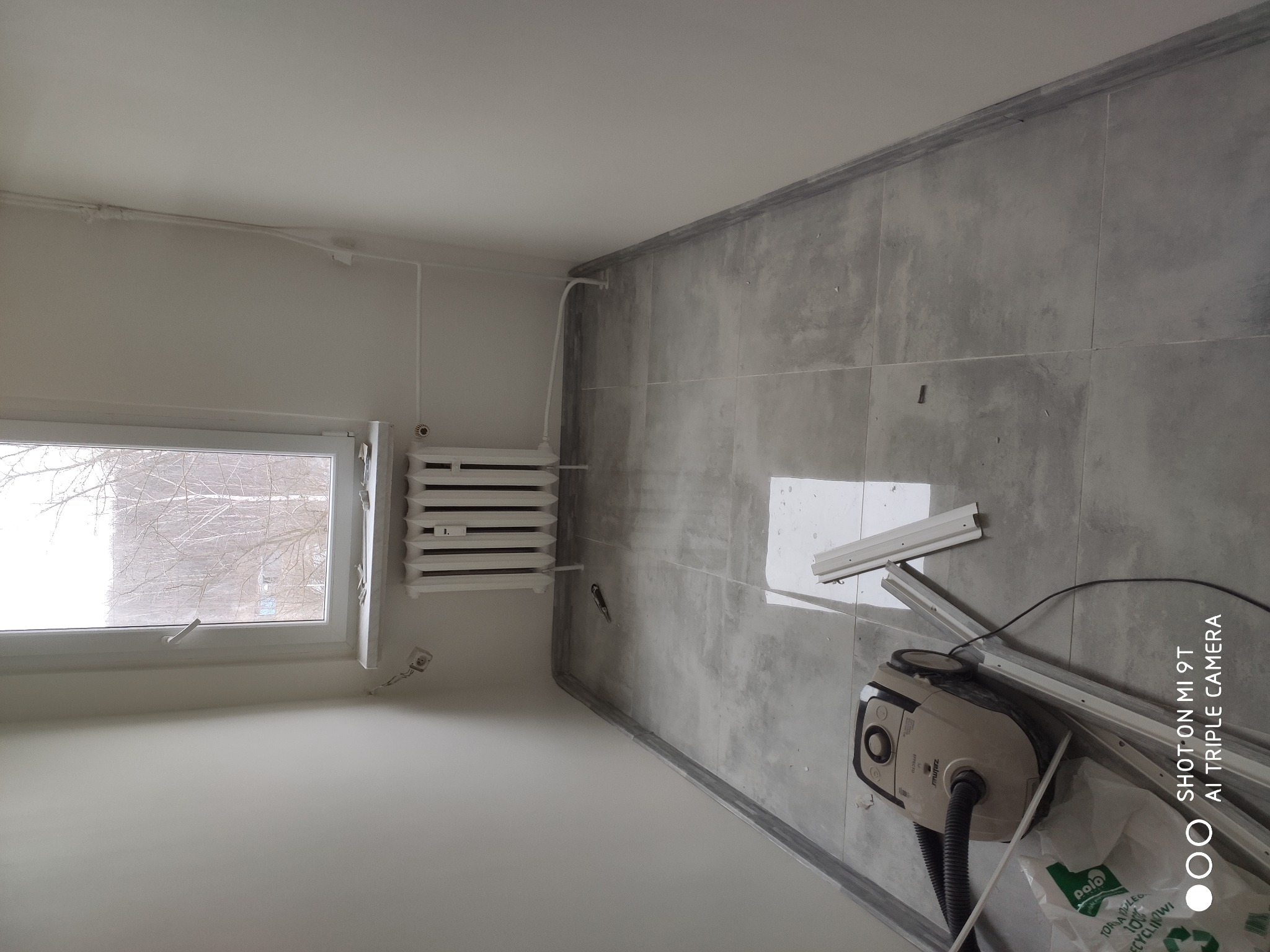 Pokój po remoncie: świeżo pomalowane białe ściany, szare płytki podłogowe, biały grzejnik pod oknem, odkurzacz i listwy przypodłogowe na podłodze.