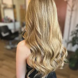 Długie, falowane włosy w odcieniach blondu z ciemniejszymi pasemkami u nasady, widok z tyłu, delikatny skręt na końcach, na ramieniu widoczny pieprzyk.