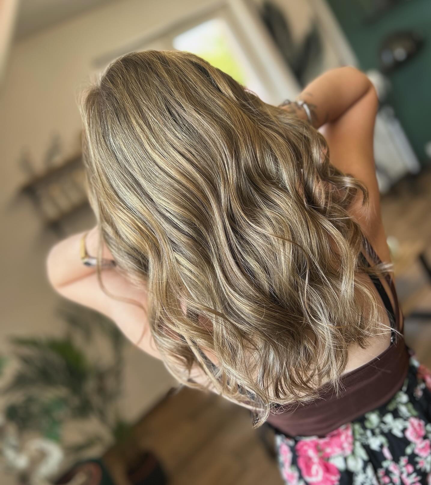 Włosy blond z refleksami, lekko falowane, widok z góry na tył głowy, na drugim planie zielona ściana i roślina doniczkowa.