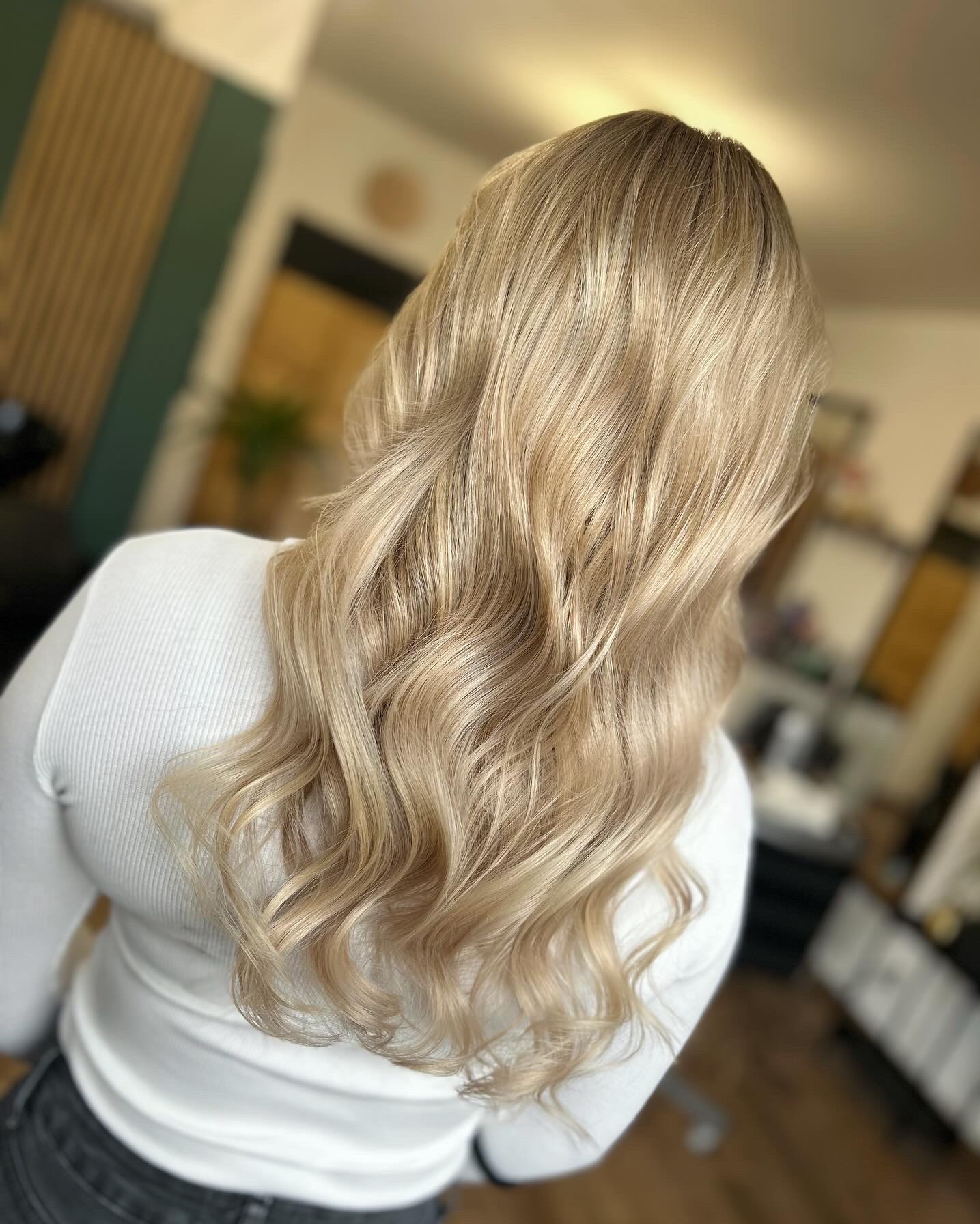Długie, blond, falowane włosy kobiety widziane od tyłu, z delikatnym balejażem, w jasnym wnętrzu salonu.