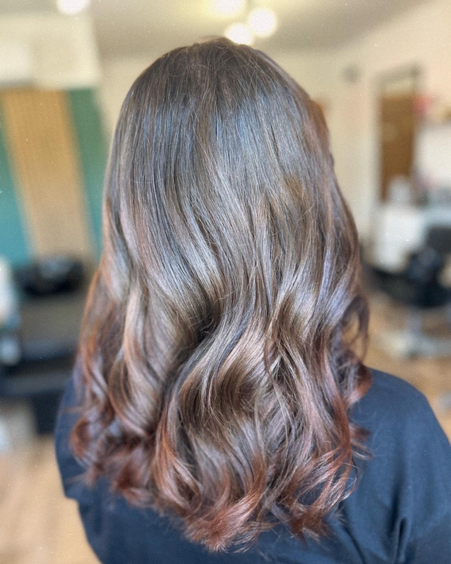 Błyszczące, brązowe włosy z delikatnymi falami, widoczne od tyłu, z subtelnymi refleksami w odcieniach miedzi na końcówkach, w tle rozmyte wnętrze salonu.