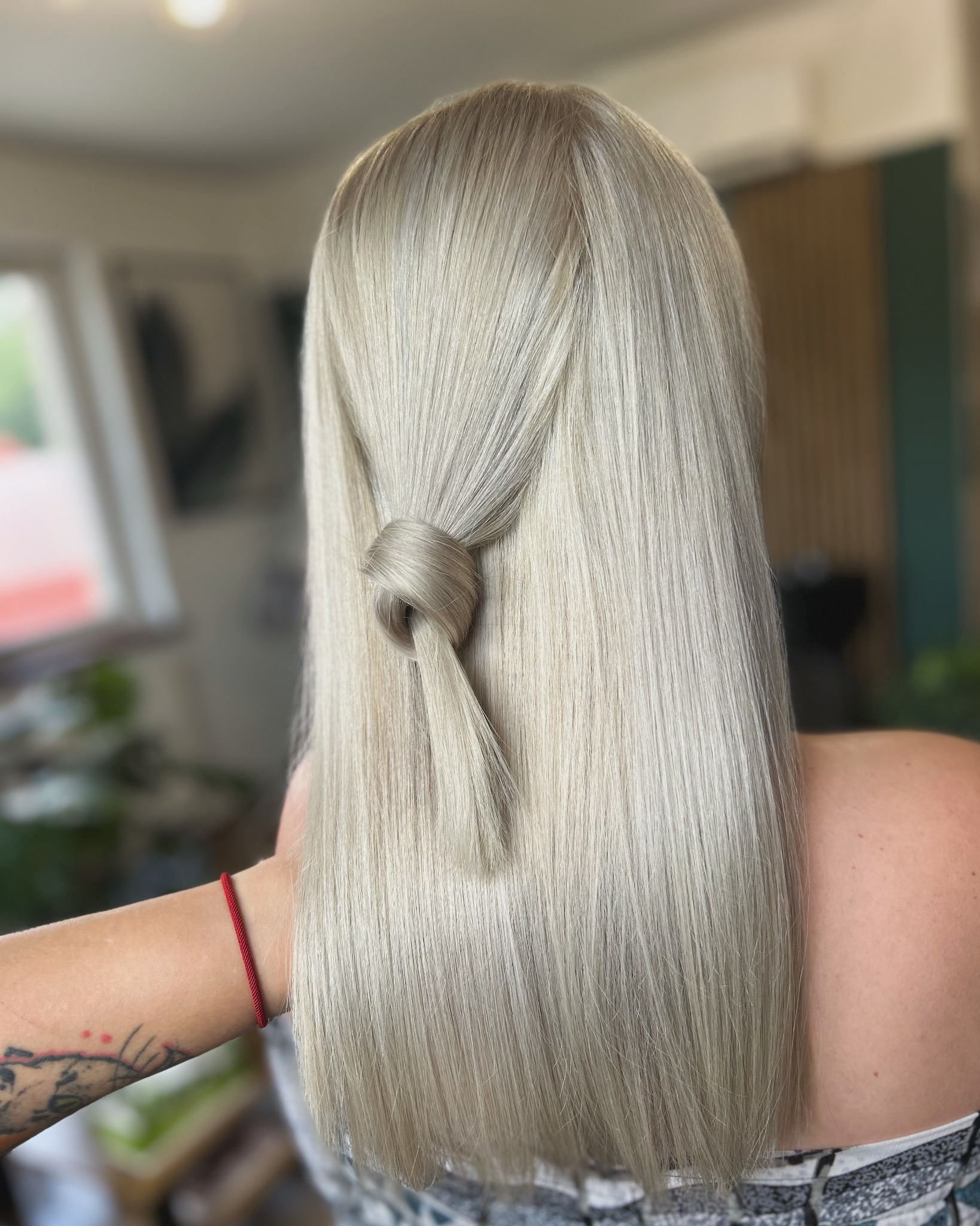 Tył głowy kobiety z długimi, platynowymi, prostymi włosami, częściowo spiętymi w supeł, widoczny tatuaż i czerwona bransoletka na ramieniu.