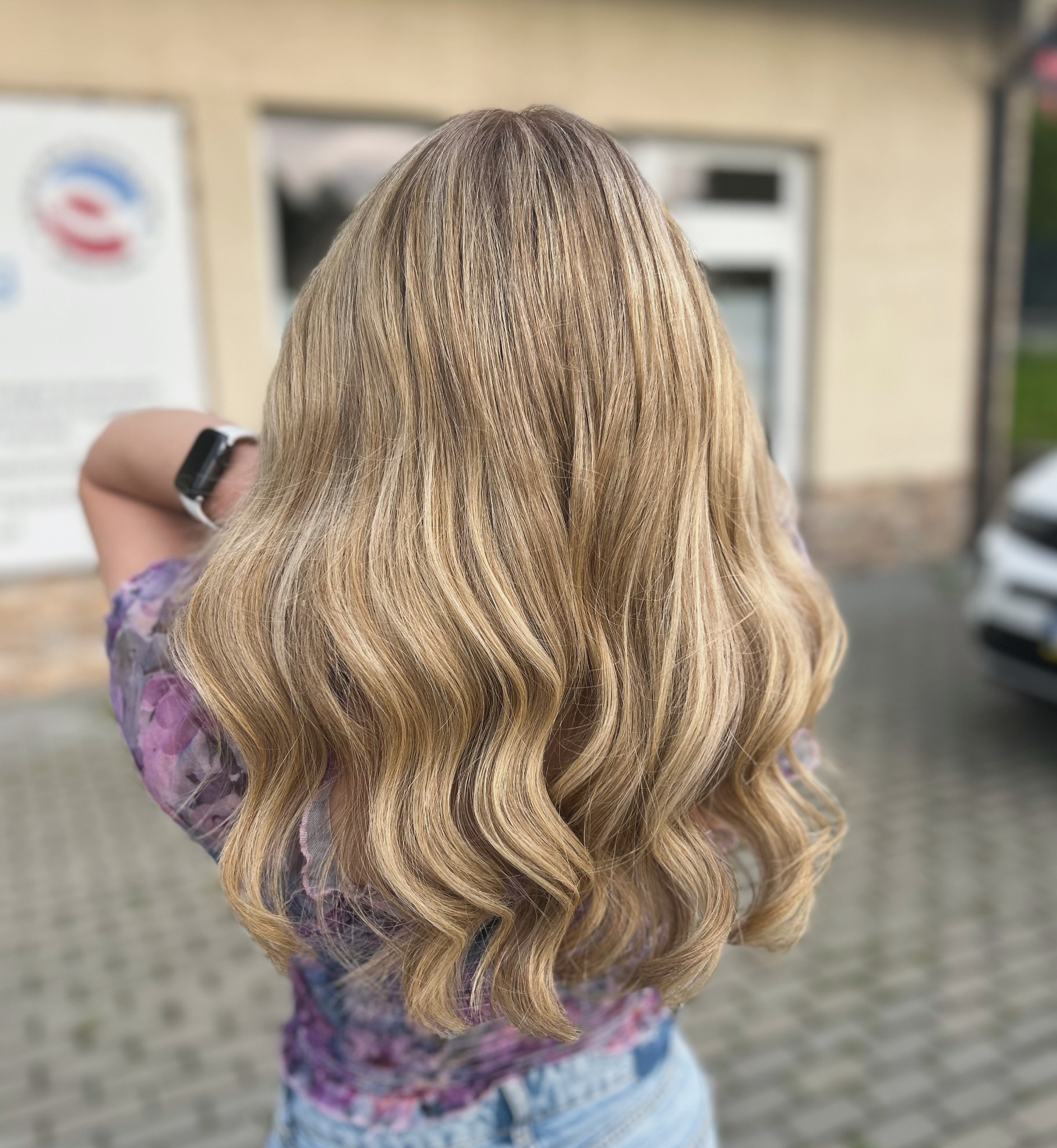 Długie, blond włosy z delikatnymi falami, widok z tyłu, z subtelnym ombre od ciemniejszych odrostów, kobieta ma na sobie bluzkę w kwiaty i jasne jeansy.