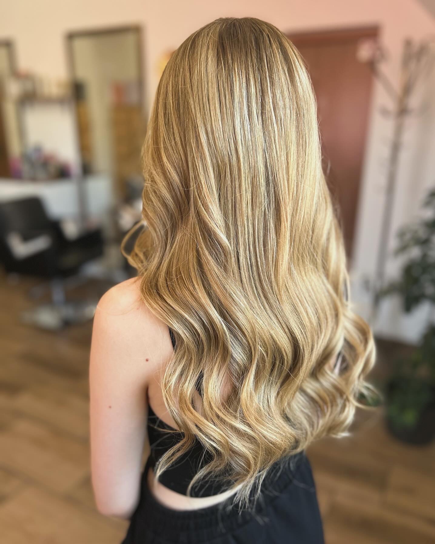 Długie, falowane włosy w odcieniach blondu z ciemniejszymi pasemkami u nasady, widok z tyłu, delikatny skręt na końcach, na ramieniu widoczny pieprzyk.