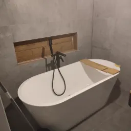 Wolnostojąca biała wanna z czarną armaturą na tle szarych płytek, z drewnianą półką na wannie i wnęką wykończoną drewnem.