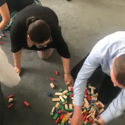 Uczestnicy szkolenia budują konstrukcje z kolorowych drewnianych klocków na szarym dywanie, ćwicząc pracę zespołową i kreatywne rozwiązywanie problemów.