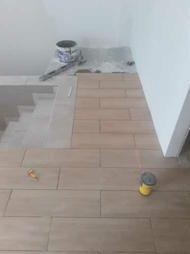 Układanie podłużnych płytek imitujących drewno na podłodze, widoczny laser krzyżowy wyznaczający linię prostą, wiadro z klejem i narzędzia w tle.