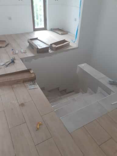 Montaż płytek podłogowych imitujących drewno na schodach i podłodze, widoczne pudełka po płytkach, narzędzia glazurnicze i fragment schodów z szarymi płytkami.