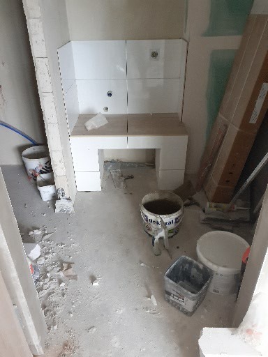 Wnętrze łazienki w trakcie prac wykończeniowych: częściowo ułożone białe płytki na ścianie, widoczne puszki instalacyjne, surowy beton na podłodze, narzędzia i pozostałości po budowie.