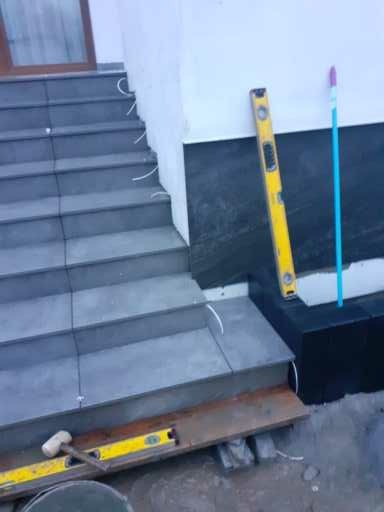 Schody zewnętrzne wyłożone szarą terakotą w trakcie prac, widoczna poziomica, gumowy młotek i niebieski mop oparty o ścianę.
