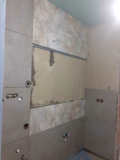 Łazienka w trakcie remontu: częściowo ułożone beżowe płytki na ścianach, widoczne otwory na instalacje, metalowa listwa montażowa i fragmenty surowej ściany.