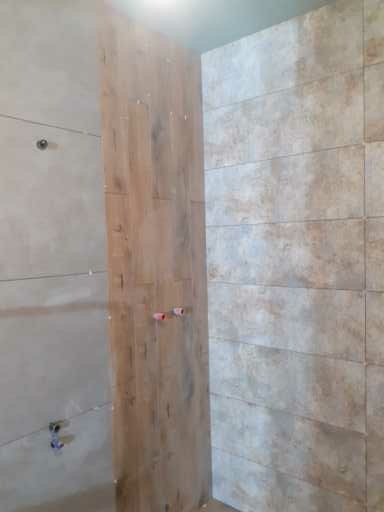 Wnętrze łazienki w trakcie prac wykończeniowych, widoczne płytki imitujące drewno na jednej ze ścian oraz jasne, beżowe płytki na pozostałych ścianach, z widocznymi otworami na armaturę.