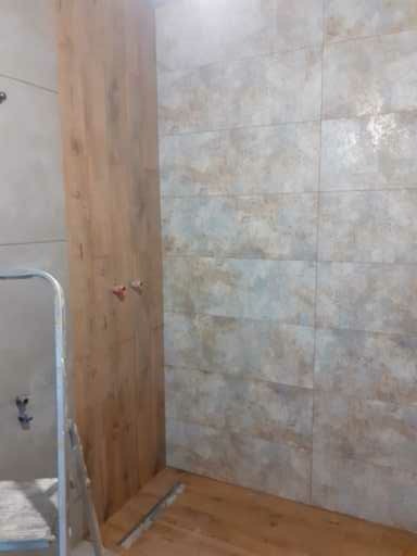 Fragment łazienki w trakcie wykańczania: ściana z płytek imitujących kamień, drewnopodobna obudowa, widoczne rury i drabinka, podłoga w trakcie układania.