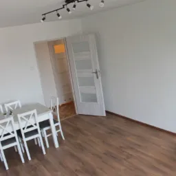 Jasne wnętrze pokoju po remoncie z drewnianą podłogą, białymi ścianami i białym stołem z krzesłami. Widoczny fragment niebieskiego pufa w rogu pokoju.