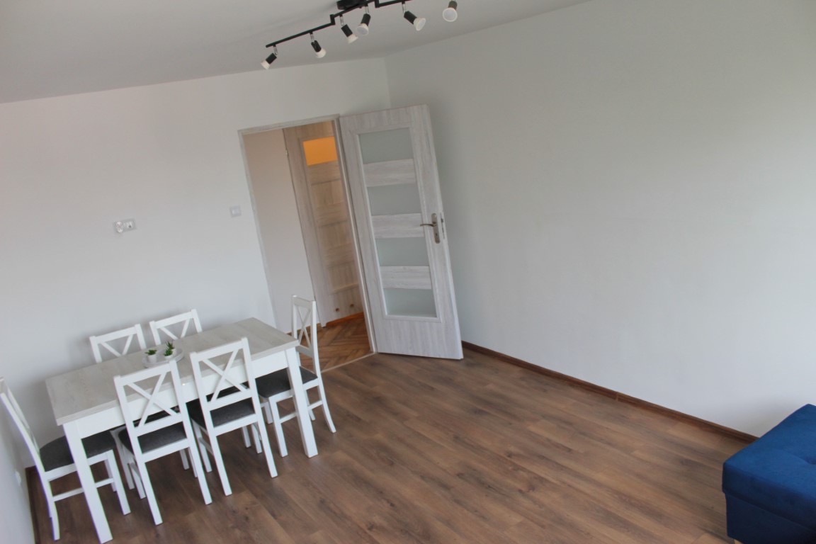 Jasne wnętrze pokoju po remoncie z drewnianą podłogą, białymi ścianami i białym stołem z krzesłami. Widoczny fragment niebieskiego pufa w rogu pokoju.