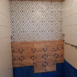 Ściana łazienki w trakcie układania płytek: górna część z białymi płytkami z geometrycznym wzorem, dolna część z płytkami imitującymi drewno, widoczne niebieskie krzyżyki dystansowe.