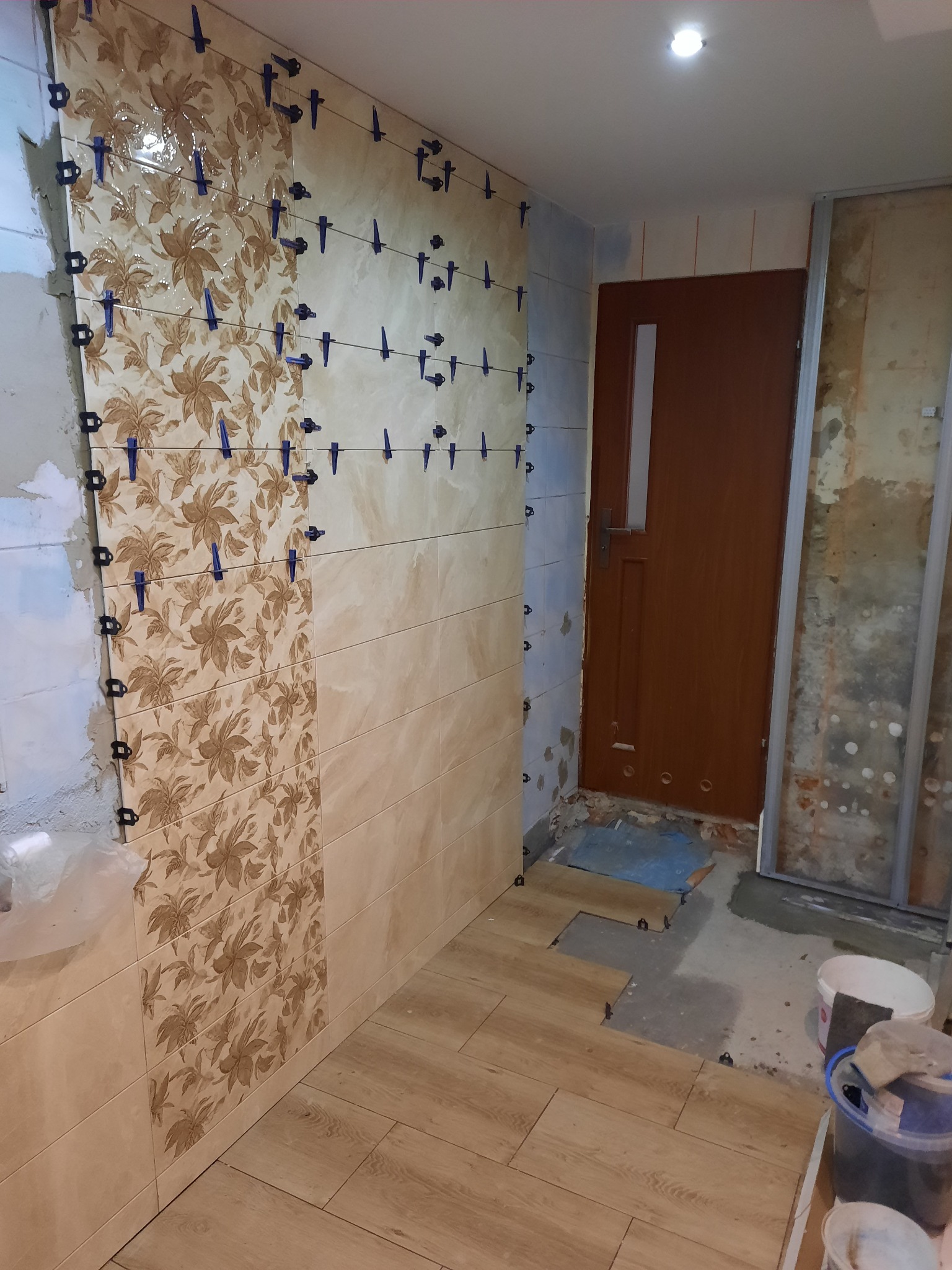 Ściana łazienki w trakcie układania płytek z wzorem kwiatowym, użyte klipsy poziomujące; podłoga częściowo wyłożona panelami imitującymi drewno, widoczna niedokończona ściana i drzwi.