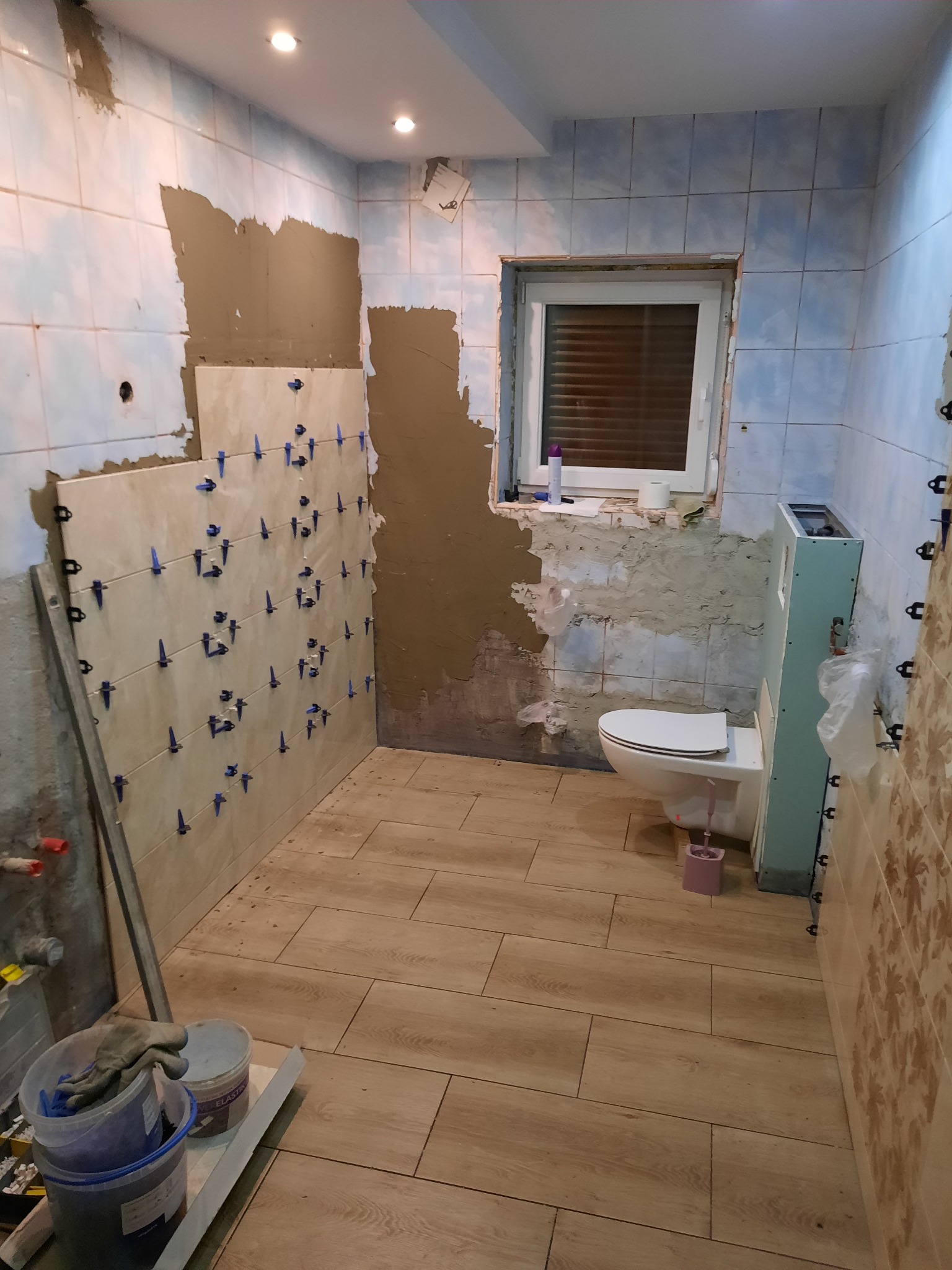 Łazienka w trakcie remontu: częściowo położone płytki na ścianach z użyciem klipsów poziomujących, przygotowane podłoże, widoczny sedes i narzędzia w wiadrach.