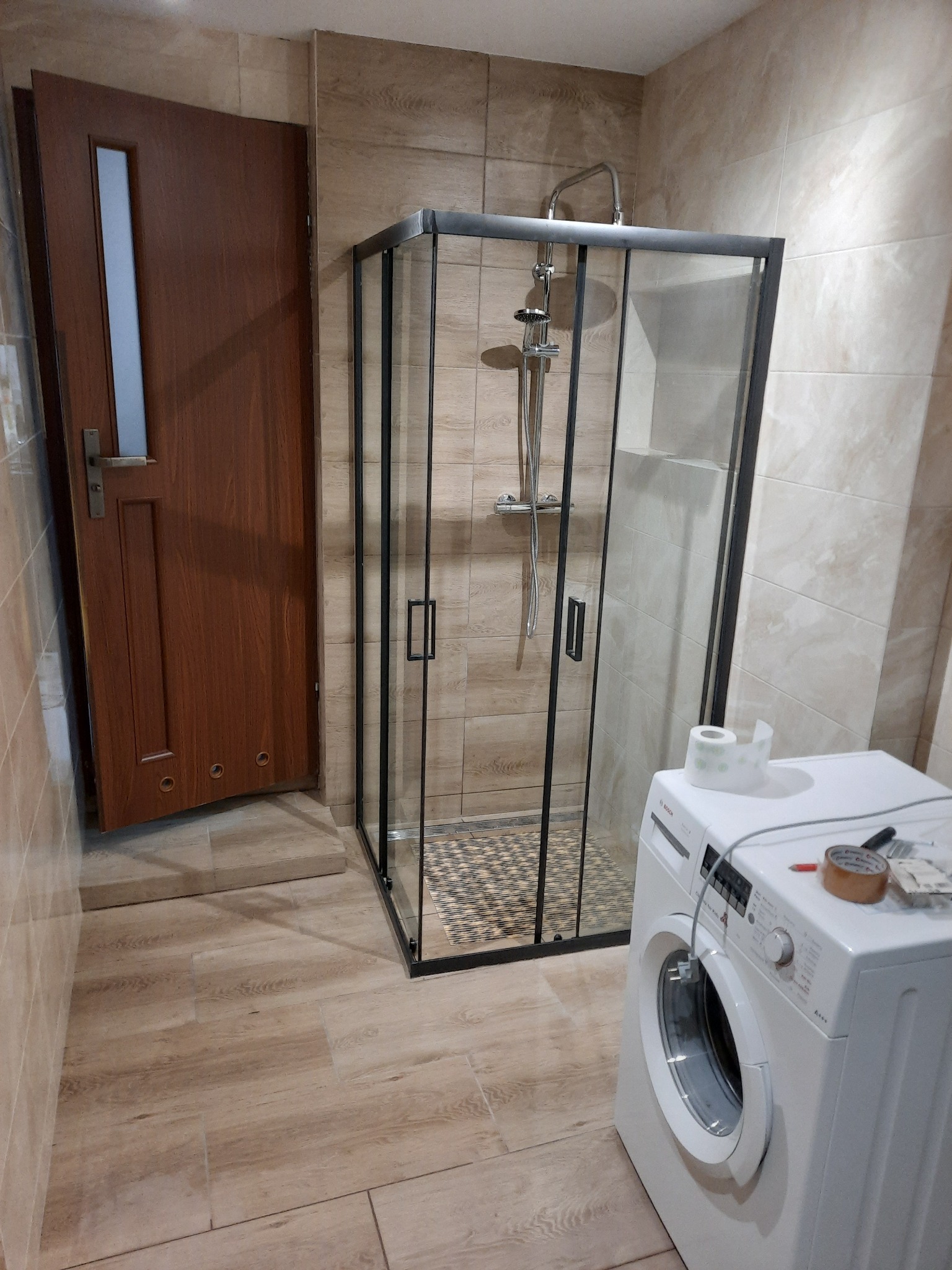 Nowoczesna kabina prysznicowa z czarnymi profilami w łazience wykończonej płytkami imitującymi drewno, obok pralka z rolką taśmy i papierem toaletowym.