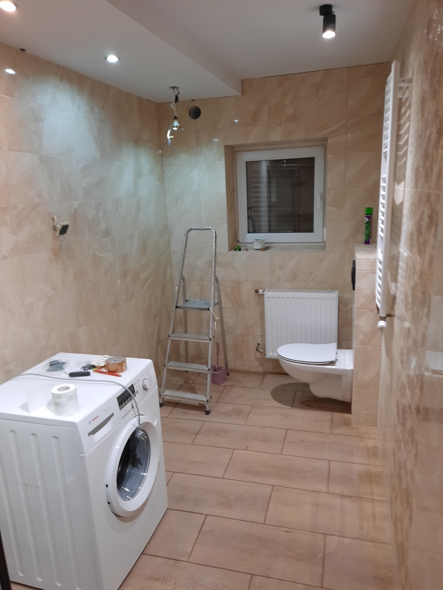 Beżowa łazienka w trakcie remontu: widoczna pralka z narzędziami na górze, drabina, toaleta, grzejnik i płytki imitujące drewno na podłodze i ścianach. Część instalacji elektrycznej jest jeszcze...