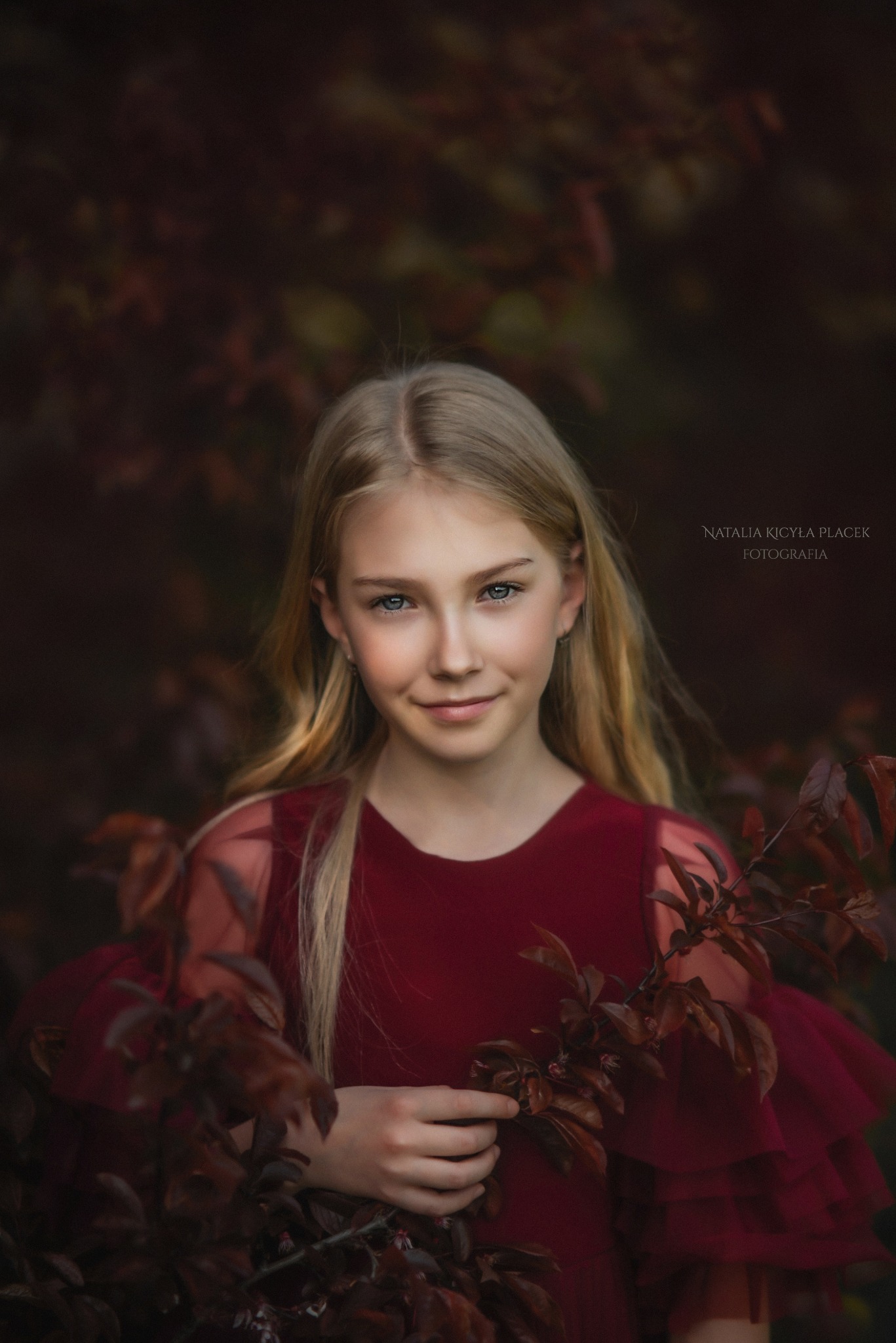 Portret uśmiechniętej blondynki w bordowej sukience na tle ciemnych liści, autorstwa Natalii Kicyły Placek. Delikatne światło, naturalny wygląd.