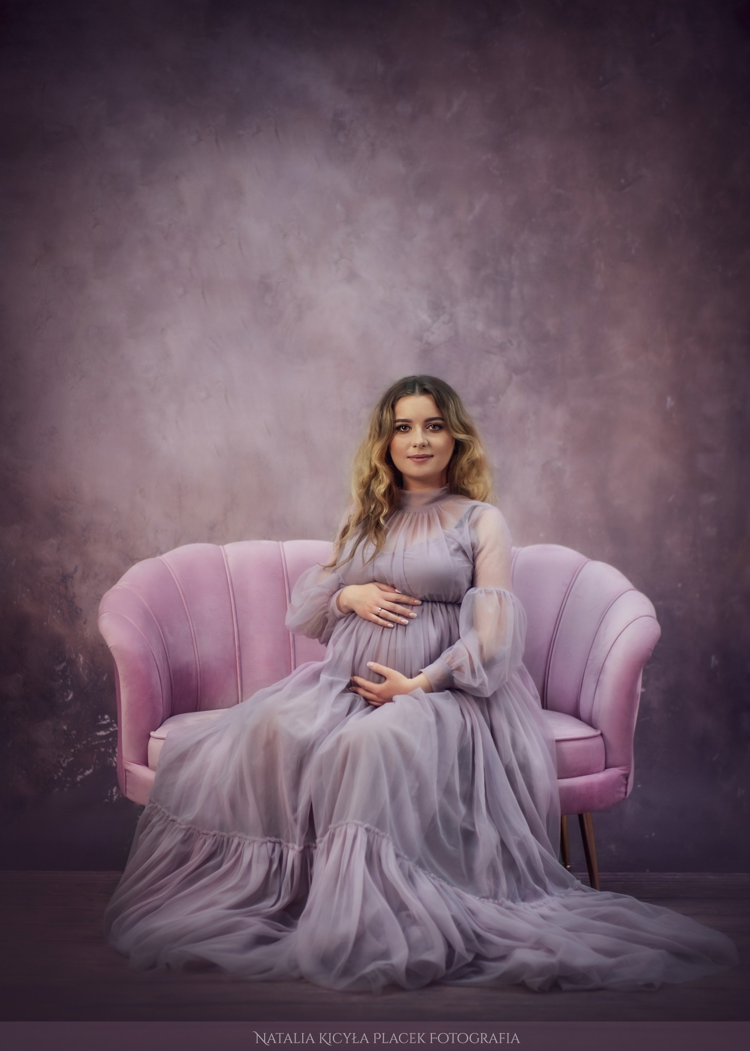 Kobieta w ciąży w długiej, zwiewnej sukni w odcieniach fioletu siedzi na różowym fotelu na tle fioletowego, teksturowanego tła. Dłonie ułożone na brzuchu.