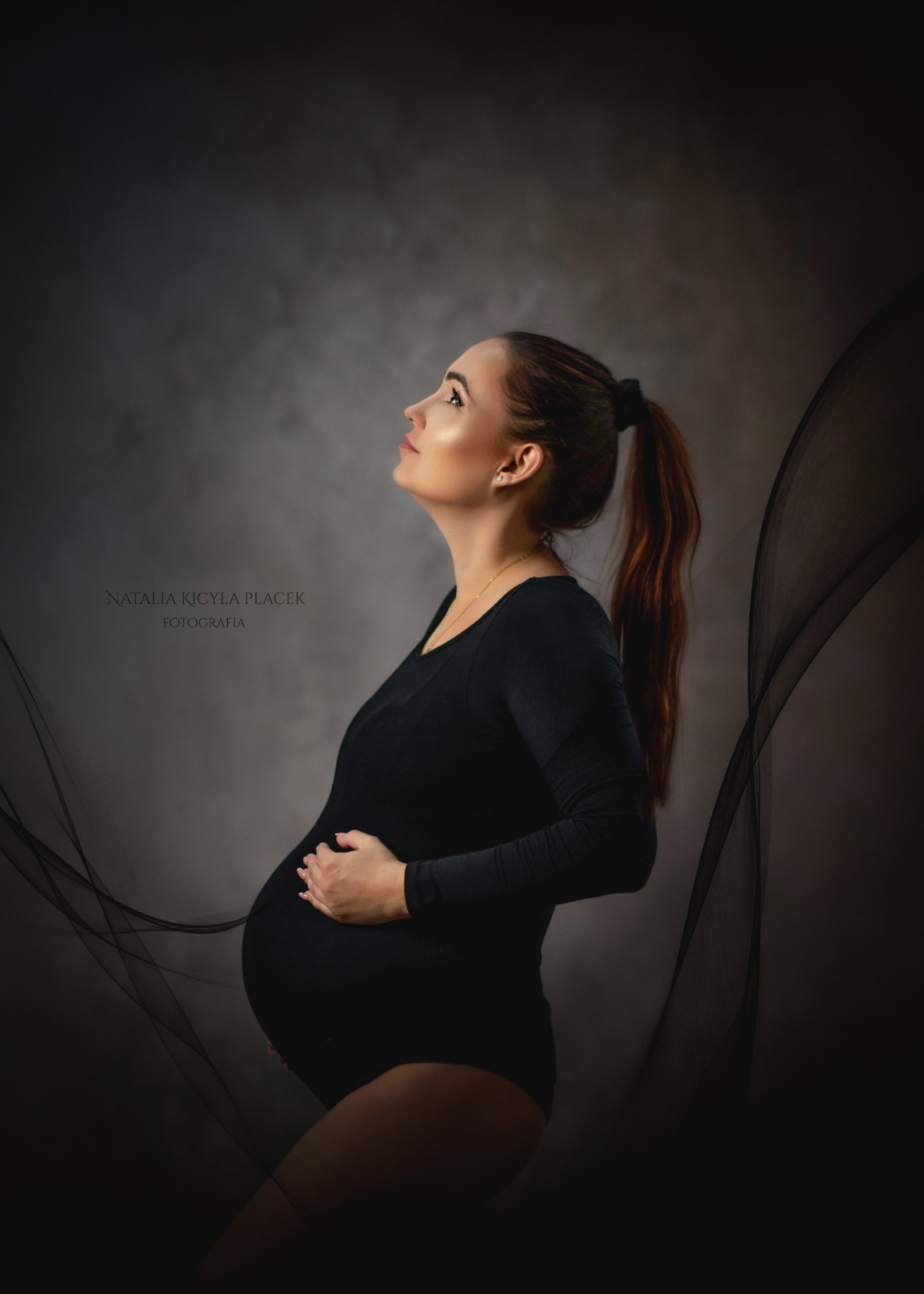 Portret kobiety w ciąży, ubrana w czarny strój, z włosami związanymi w kucyk, spogląda w górę na tle szarego, abstrakcyjnego tła z czarnymi, płynącymi liniami.