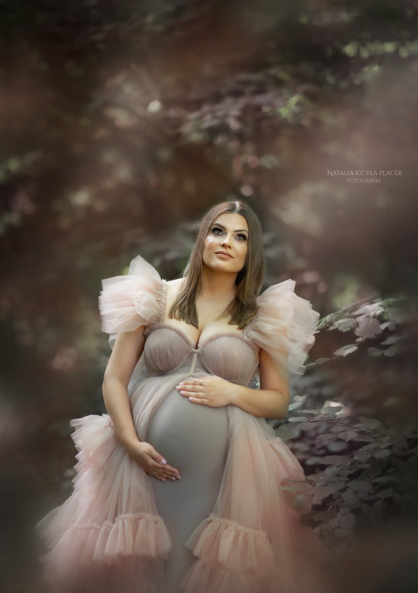 Kobieta w ciąży w tiulowej sukni z falbanami, pozująca na zewnątrz z rękami na brzuchu, spoglądająca w górę w miękkim świetle.