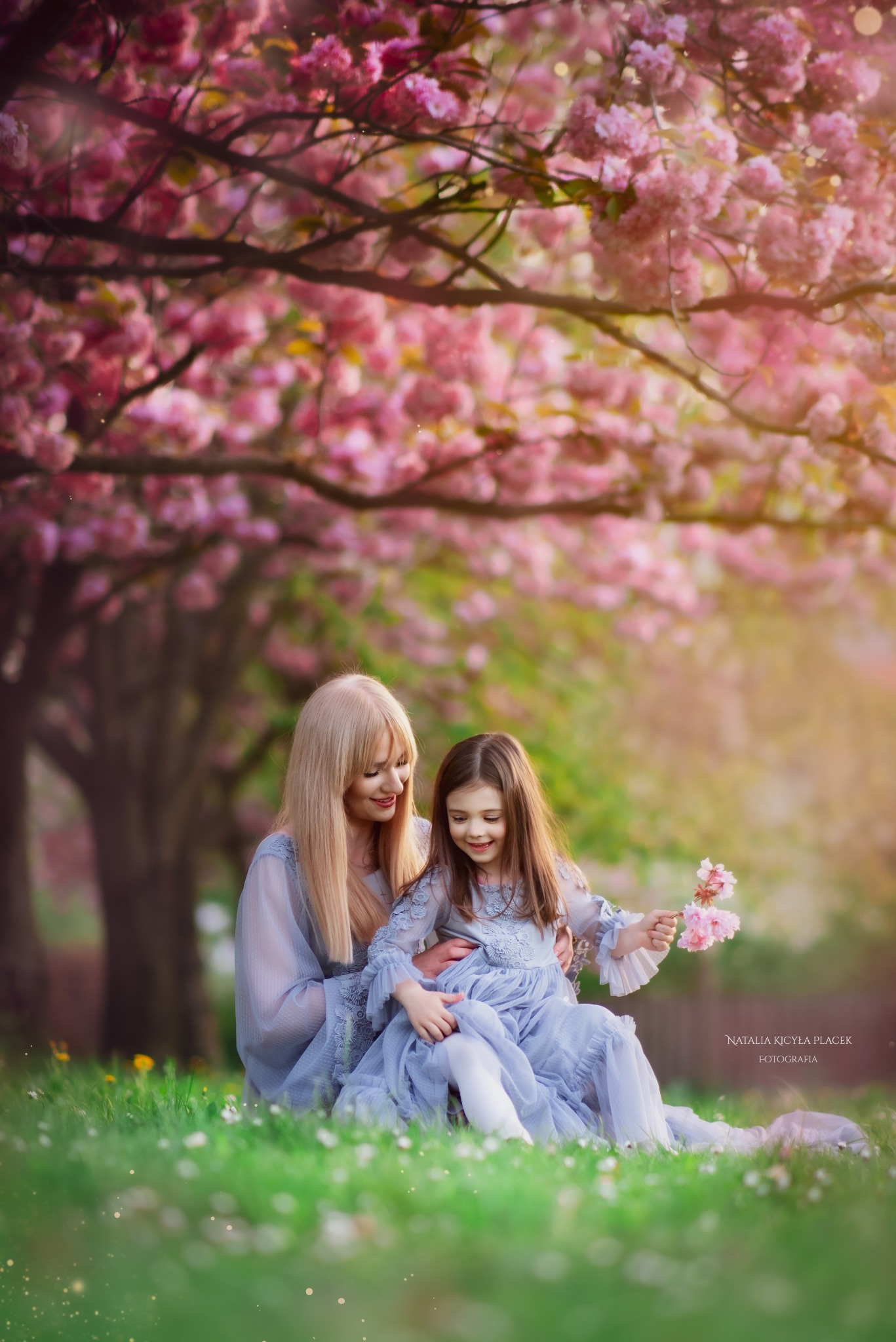 Matka z córką w dopasowanych, błękitnych sukienkach siedzą na trawie pod kwitnącą wiśnią, trzymając w dłoniach gałązki kwiatów.