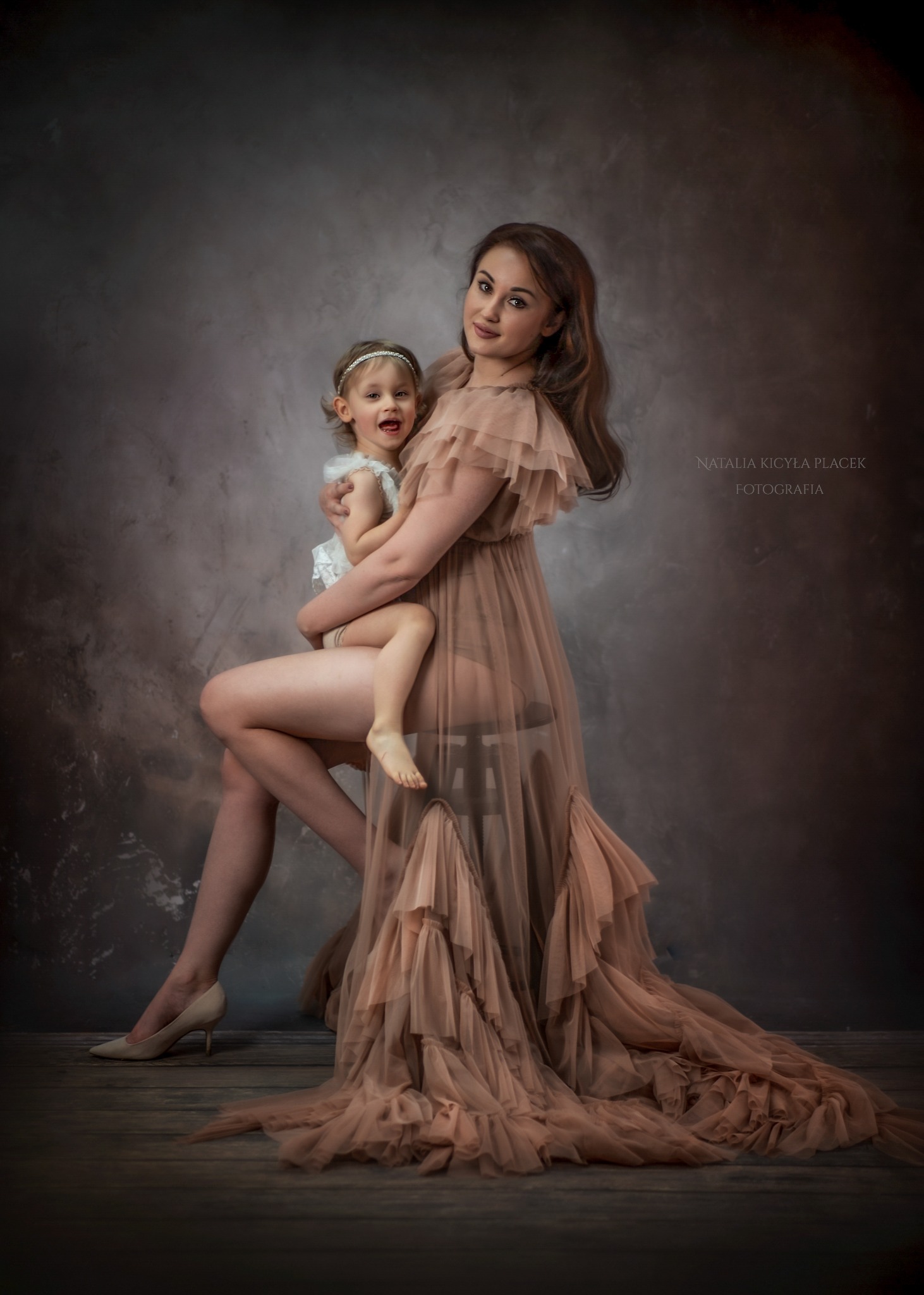 Elegancka kobieta w zwiewnej, warstwowej sukni w kolorze nude, siedząca z małą dziewczynką na kolanach, na tle ciemnego, teksturowanego tła. Cieliste szpilki na nogach kobiety.