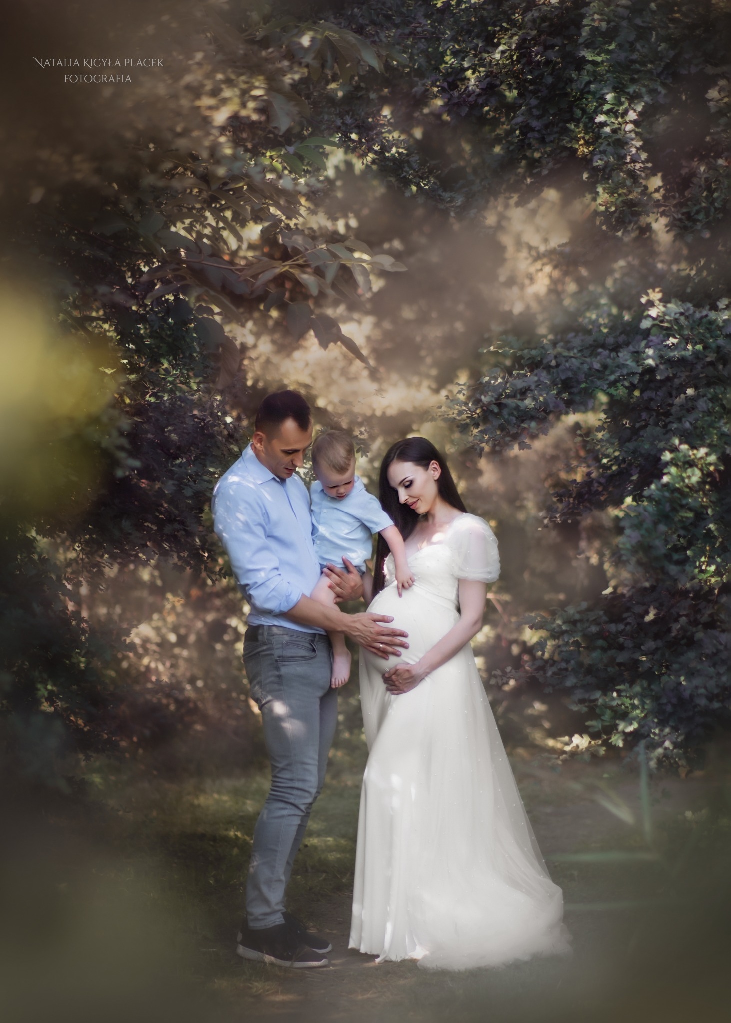 Rodzinne zdjęcie w plenerze: ojciec trzyma synka, matka w białej sukni ciążowej, oboje dłońmi obejmują brzuch, otoczeni zielenią.