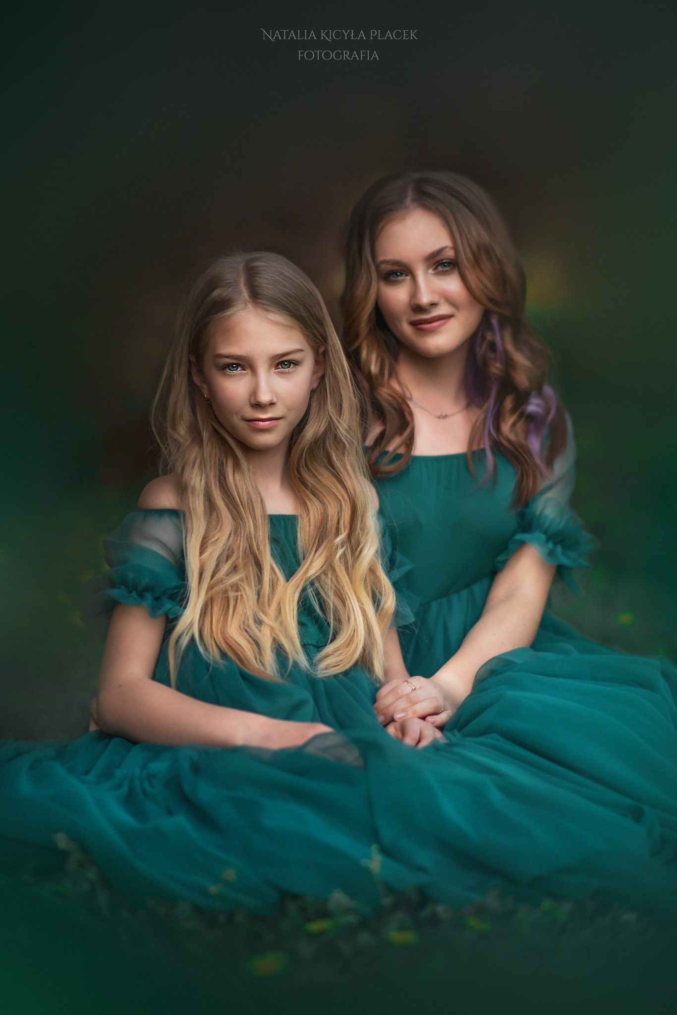 Portret matki i córki w szmaragdowych sukienkach, siedzących blisko siebie na tle ciemnej zieleni, z delikatnym oświetleniem podkreślającym ich rysy twarzy i długie włosy.