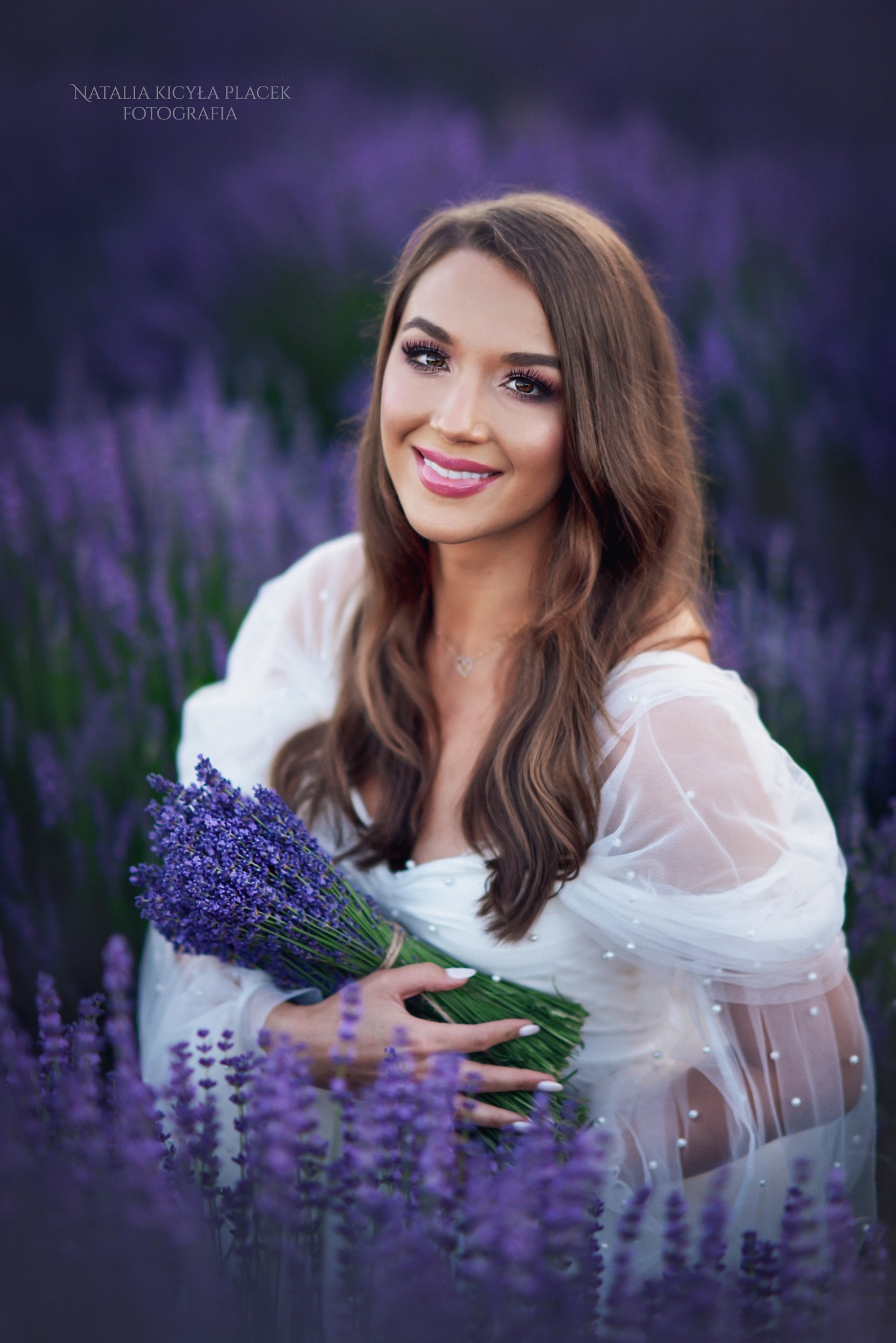 Uśmiechnięta kobieta w zwiewnej, białej sukience z perłami, trzyma bukiet lawendy na tle kwitnącego pola lawendowego.