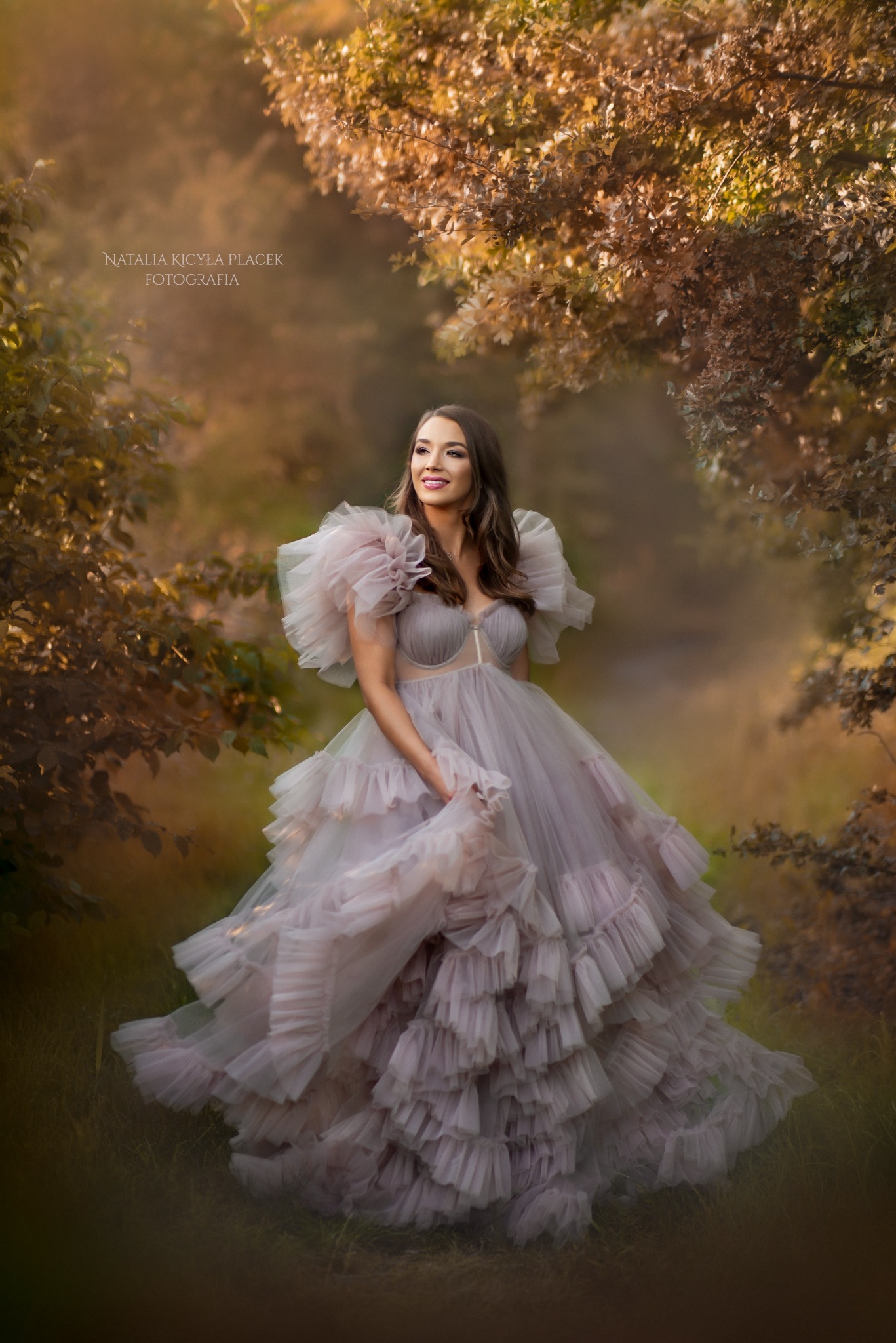 Uśmiechnięta kobieta w tiulowej sukni z falbanami, w plenerze w okolicach Opola, pozuje na tle drzew z jesiennymi liśćmi.