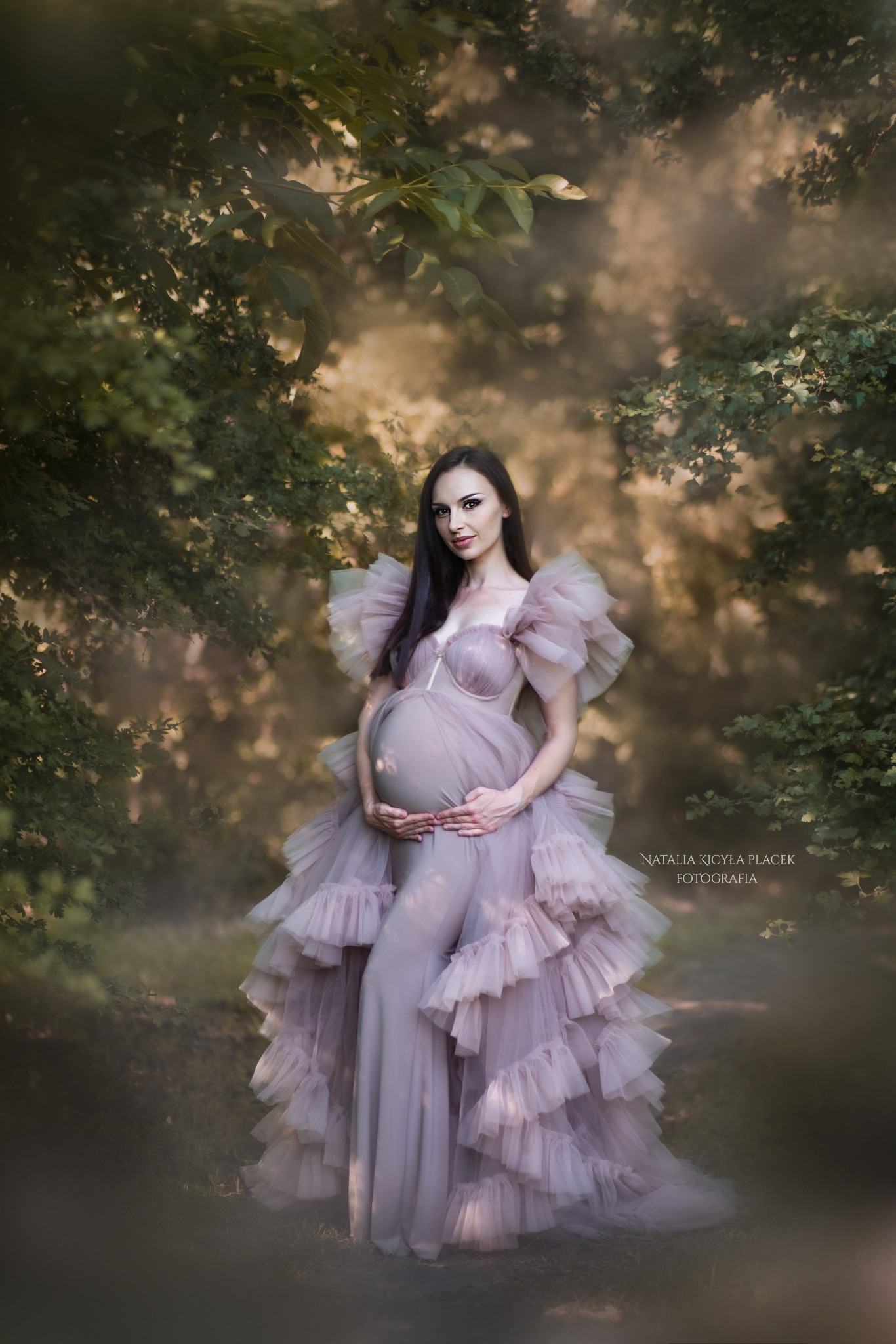 Kobieta w ciąży w długiej, warstwowej sukni z falbanami w odcieniach pudrowego różu, pozująca w leśnej scenerii z delikatnym, rozproszonym światłem.