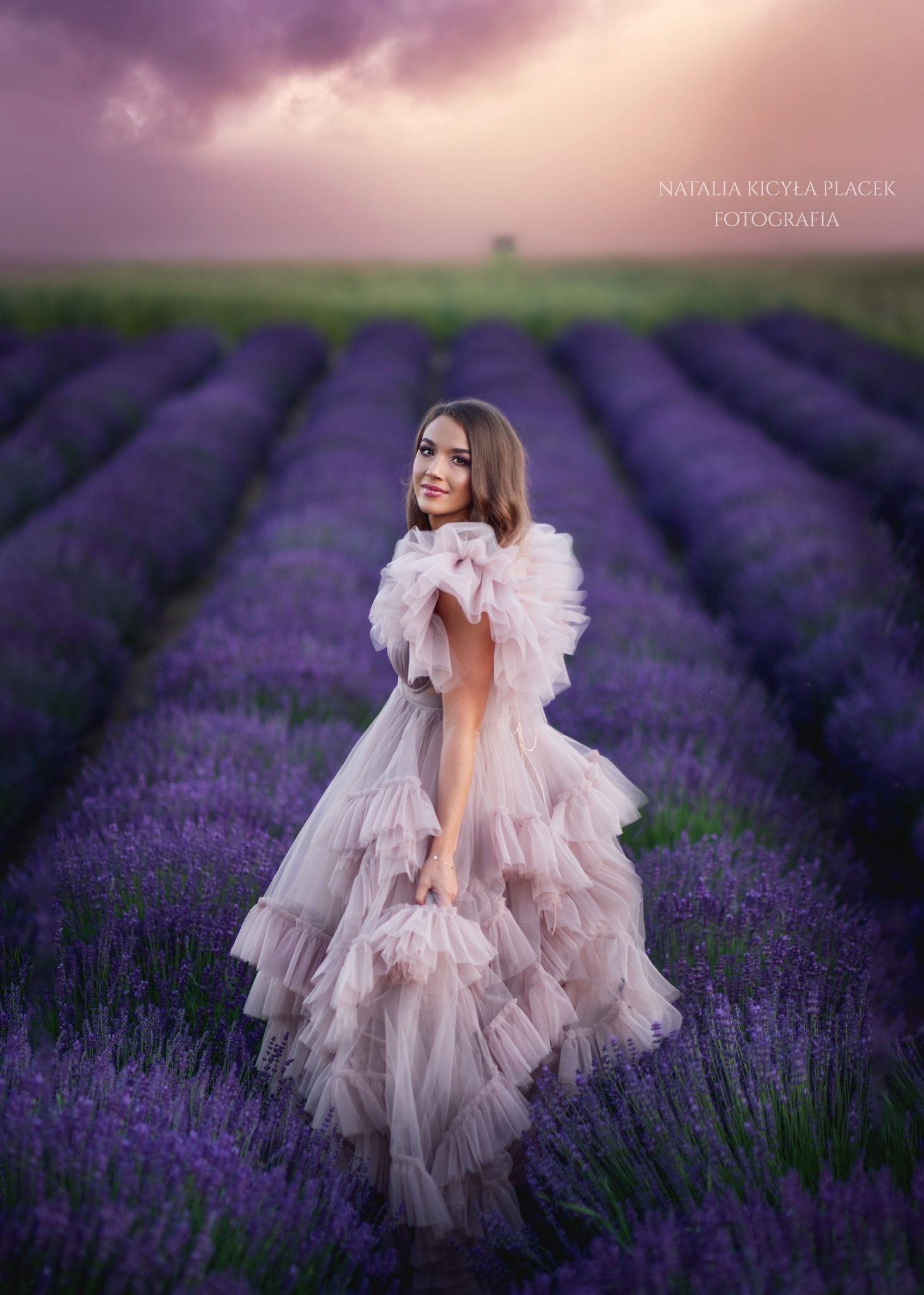 Kobieta w tiulowej sukni pozuje na tle pola lawendy o zachodzie słońca, spoglądając przez ramię.