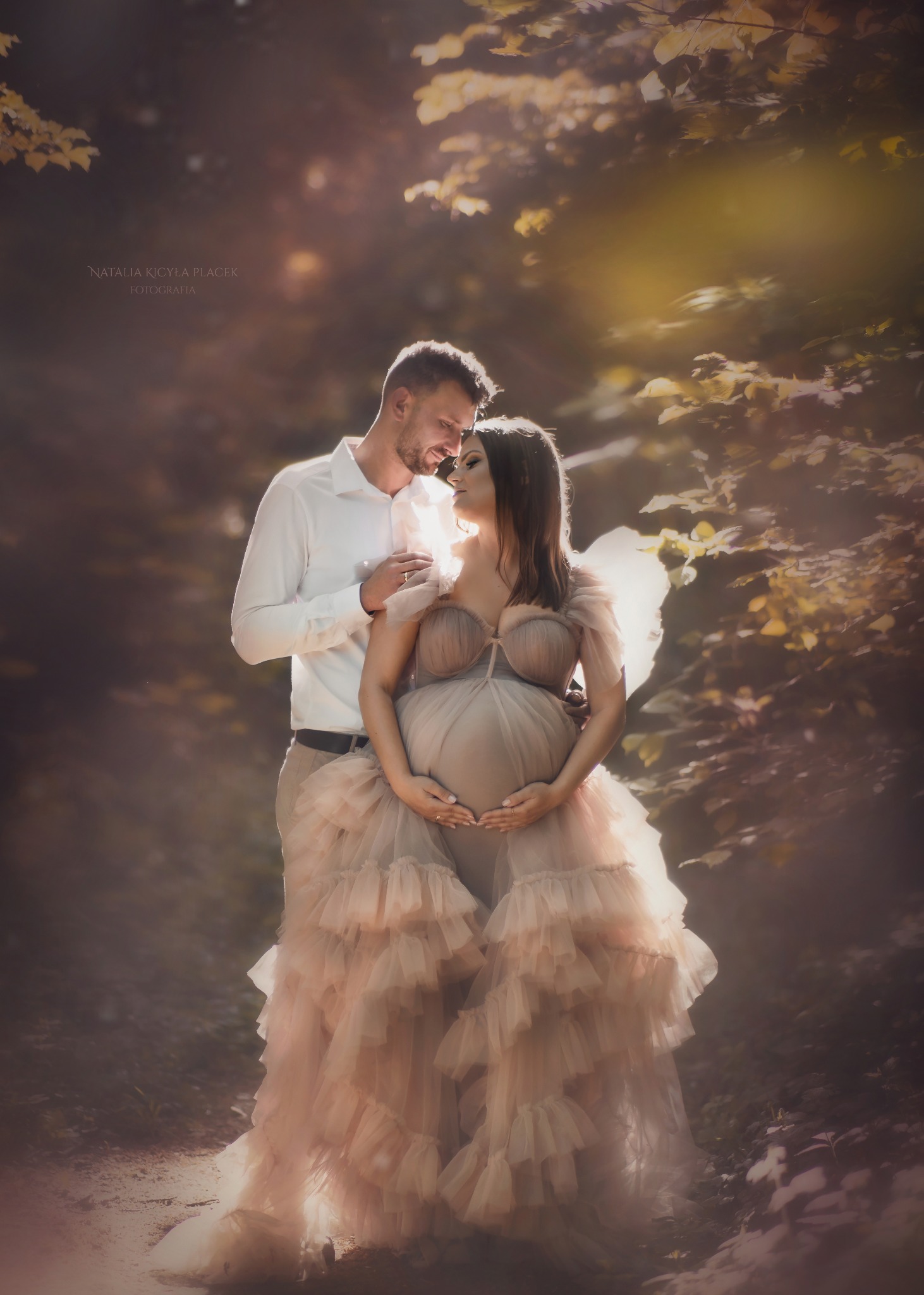 Kobieta w beżowej, warstwowej sukni ciążowej, trzymająca ręce na brzuchu, stoi z mężczyzną w białej koszuli w leśnej scenerii z promieniami słońca.