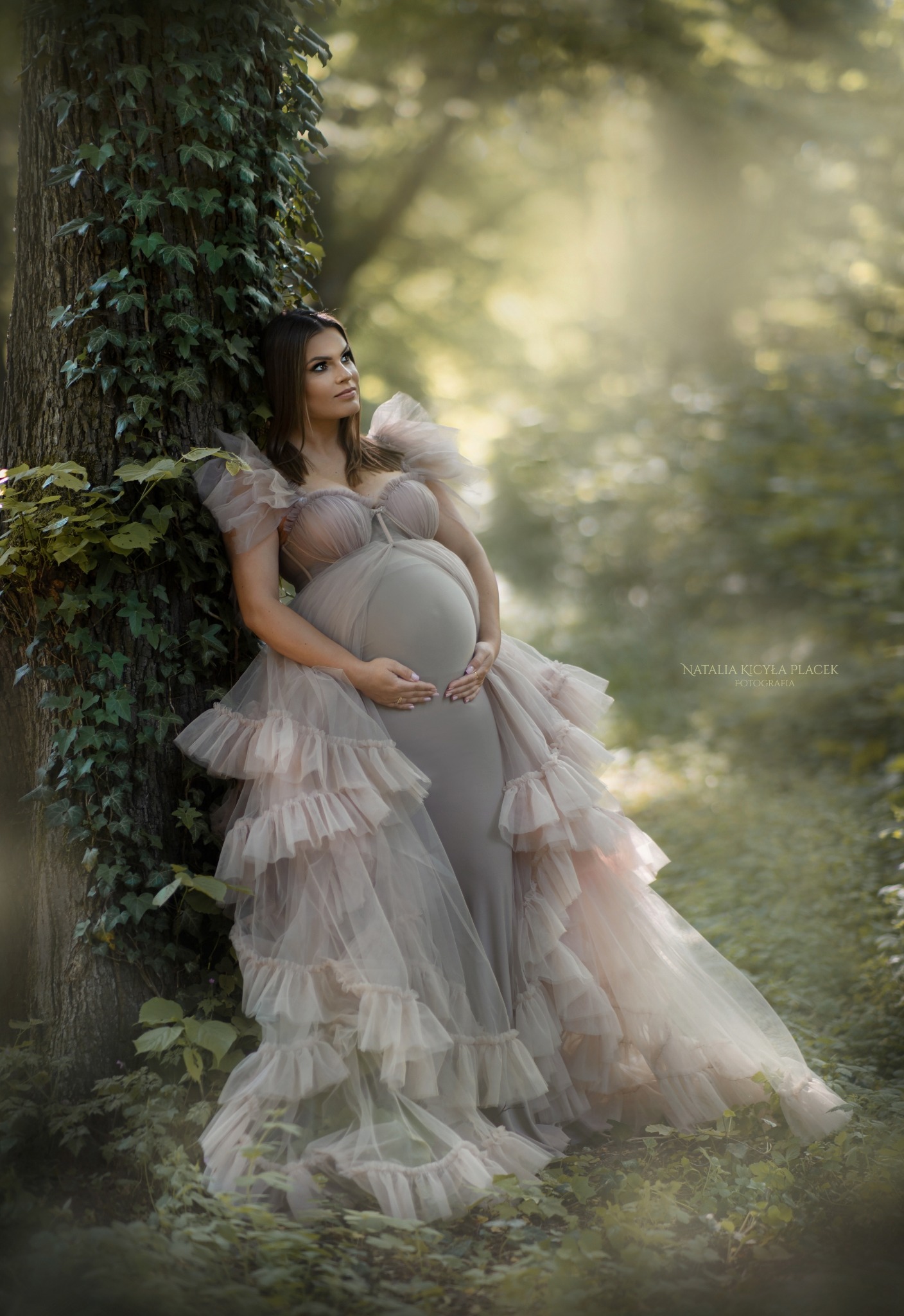 Kobieta w zwiewnej sukni, opierająca się o drzewo porośnięte bluszczem w leśnej scenerii, delikatne światło podkreśla ciążowy brzuszek.