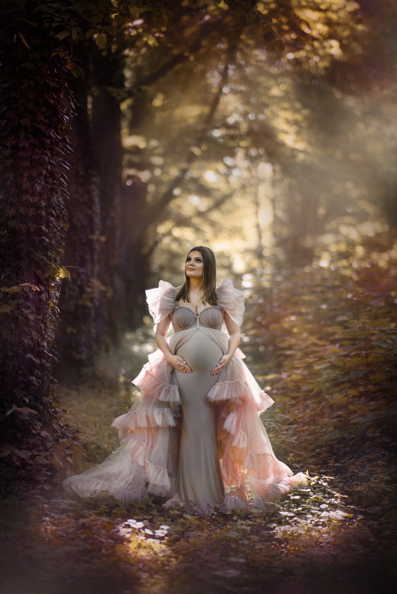 Kobieta w zaawansowanej ciąży, ubrana w elegancką, długą suknię z falbanami, pozuje w leśnej scenerii z promieniami słońca przebijającymi przez korony drzew.