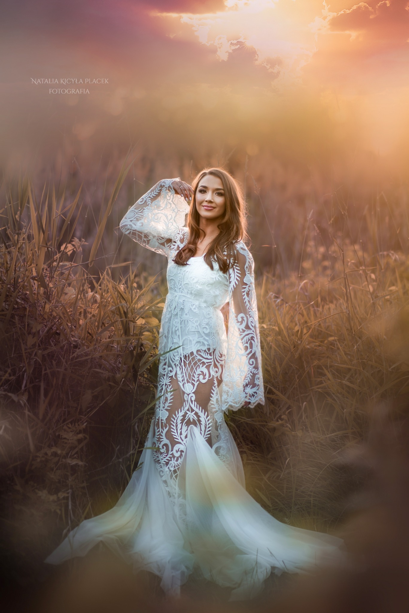 Kobieta w koronkowej sukience pozuje na tle łąki w promieniach zachodzącego słońca, z delikatnym uśmiechem i ręką uniesioną do góry.