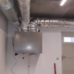 Szara jednostka centralna wentylacyjna z widocznymi przewodami wentylacyjnymi z izolacją aluminiową pod sufitem w pomieszczeniu z białą ścianą i drzwiami.