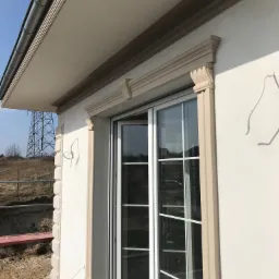 Wykończona elewacja budynku z dekoracyjnymi elementami sztukaterii wokół okna przesuwnego; widoczny fragment dachu z rynną i słup wysokiego napięcia w tle.