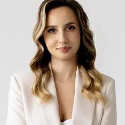 Portret kobiety z długimi, falowanymi włosami w beżowym żakiecie, uśmiechającej się do kamery na jasnym tle. Profesjonalny wizerunek.