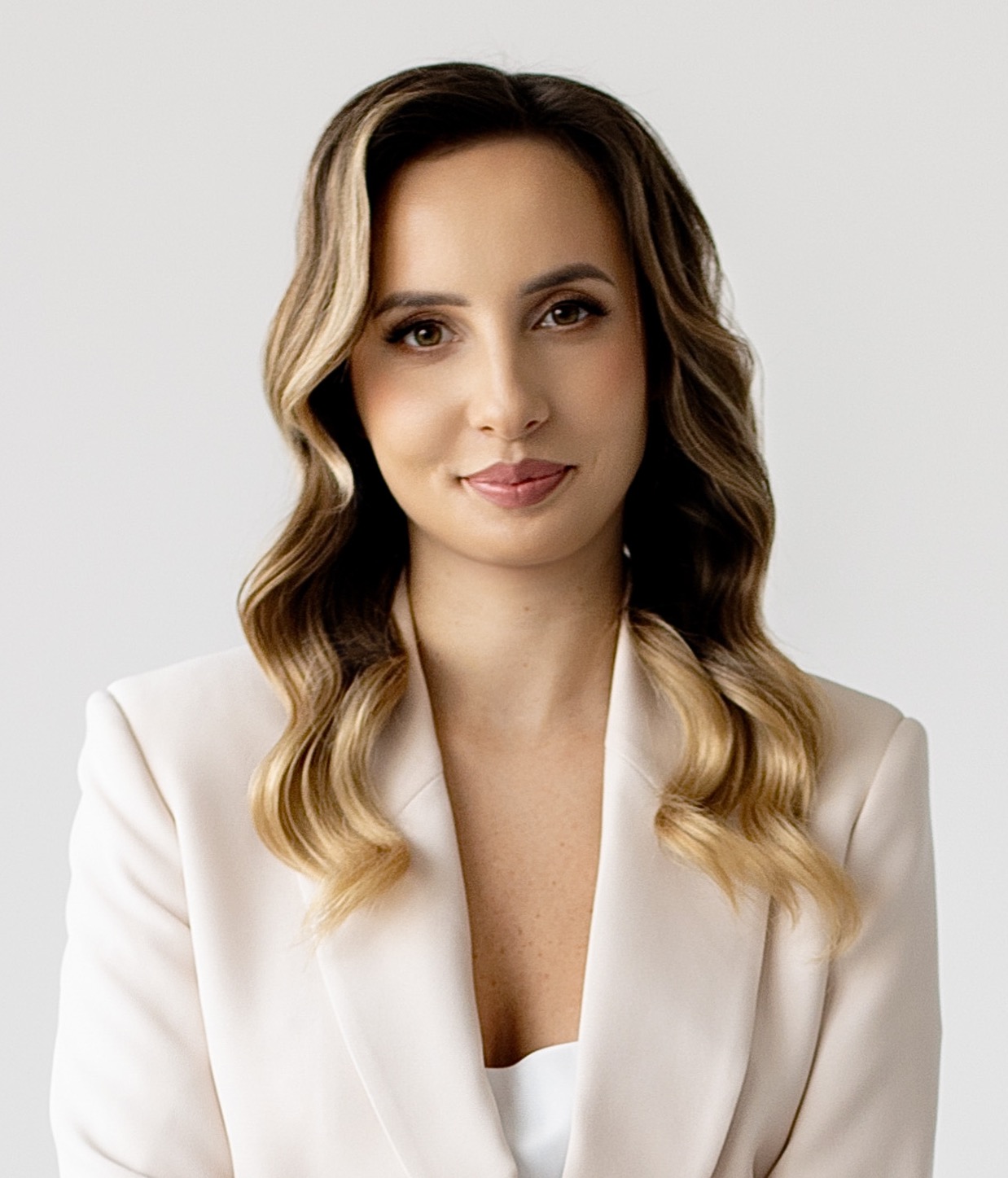 Portret kobiety z długimi, falowanymi włosami w beżowym żakiecie, uśmiechającej się do kamery na jasnym tle. Profesjonalny wizerunek.