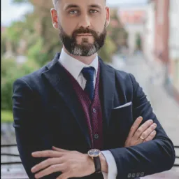 Portret mężczyzny w garniturze z założonymi rękami, na tle rozmytej przestrzeni miejskiej z zielenią.