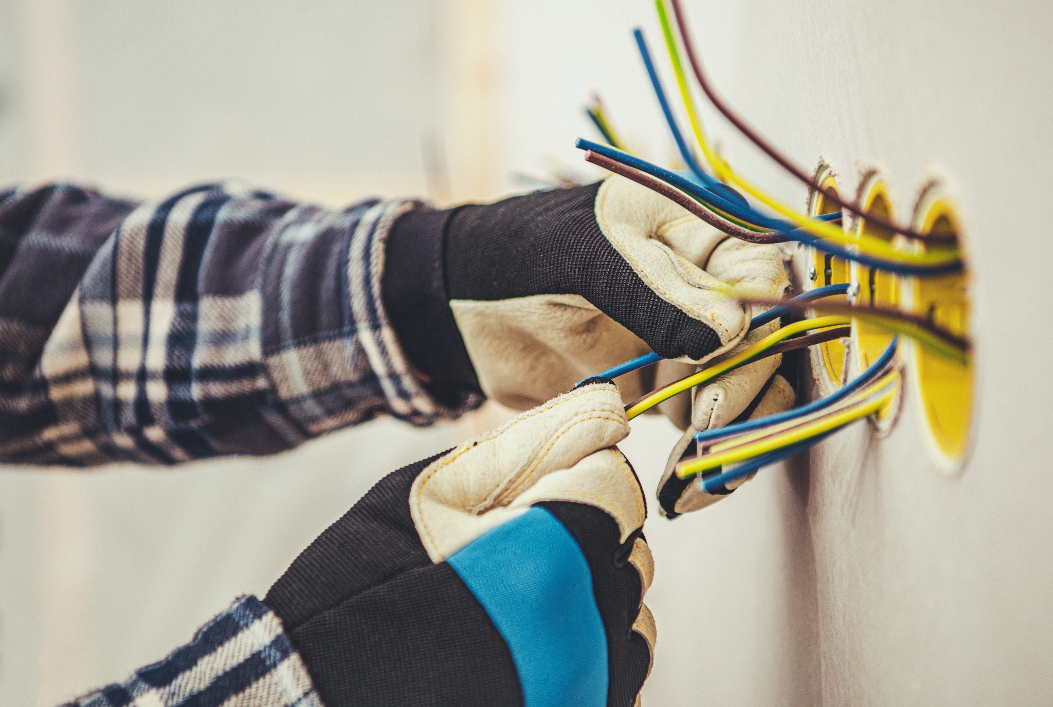 Instalacja przewodów elektrycznych w gniazdach podtynkowych, widoczne dłonie w roboczych rękawicach, przewody w izolacji żółtej, niebieskiej i brązowej, ściana w tle.
